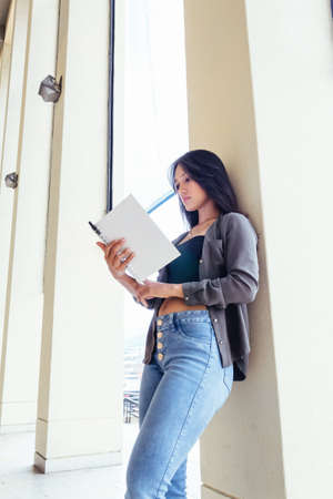 beautiful latin teen student woman with notebook, book, reading and studying leaning on school wall, learning outdoors, back to school.の写真素材