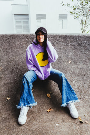 beautiful teen girl in sweatshirt and cap sitting in the city in urban style posing to the camera,...