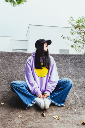 beautiful teen girl in sweatshirt and cap sitting in the city in urban style posing to the camera, happy trendy latin girl outdoorsの写真素材