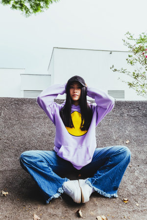 beautiful teen girl in sweatshirt and cap sitting in the city in urban style posing to the camera,...