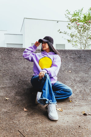 beautiful teen girl in sweatshirt and cap sitting in the city in urban style posing to the camera, happy trendy latin girl outdoorsの写真素材