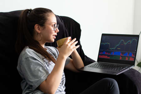 Woman drinking coffee while viewing stock and investment charts on her laptop.の写真素材