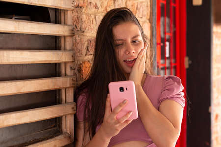 Surprised woman looks at her pastel pink cell phone, wearing pastel pink blouseの写真素材