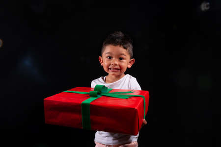 Smiling boy giving a Christmas present to the camera. On black background. Latino child isolated.の写真素材