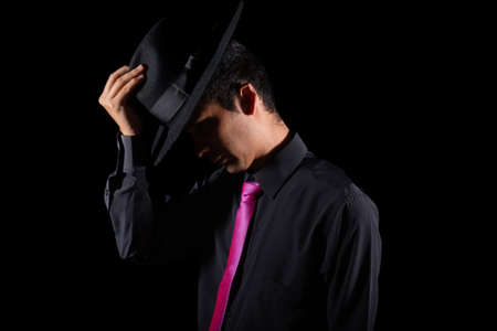 Elegant man putting on a hat in the dark, isolated on black background. Young adult young man seen in profile wearing fancy clothes.の写真素材