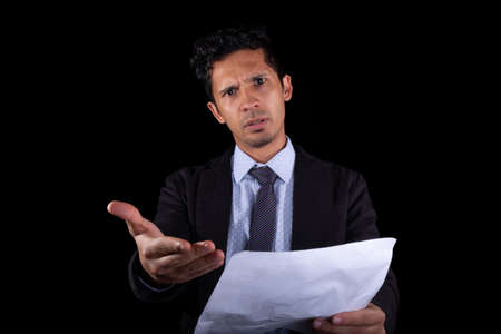Businessman making paperwork claim and looking at the camera very angry. Young businessman showing paper that was not signed, isolated on black background.の写真素材