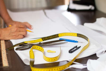 Dressmaking accessories on a fabric cutting table next to white fabric and background hands of a...