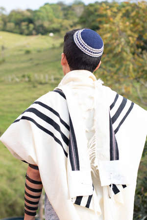 Vertical photo. Jewish man praying with talit, kippah and tefillin in nature with beautiful green meadow in the background.の写真素材