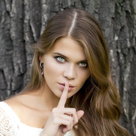 Young beautiful brunette woman has put forefinger to lips as sign of silence, against green summer gardenの写真素材