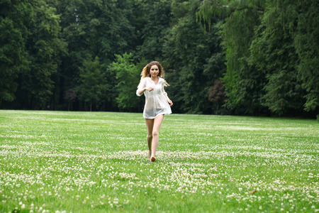 Young beautiful girl in the white shirt is running on the green field in summer parkの写真素材