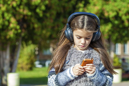 Beautiful little girl listening to music on headphones in autumn parkの写真素材