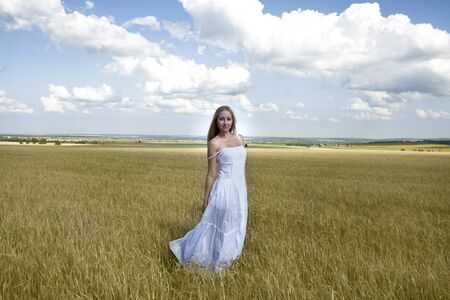 Full length portrait of Young beautiful woman in a long white dress is standing in a wheat fieldの写真素材