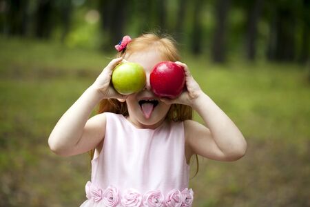 Happy little red-haired girl in a pink dress holding two apples, summer green parkの写真素材