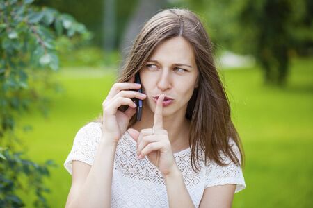 Young beautiful brunette woman has put forefinger to lips as sign of silence, against green summer...