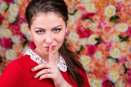Young beautiful brunette woman has put forefinger to lips as sign of silence in a studio on a floral...