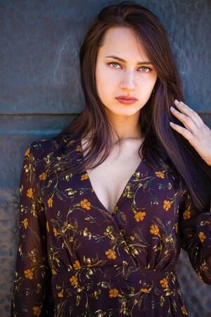 Beautiful young brunette woman in sexy brown dress, against still wall, summer outdoors