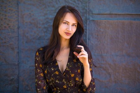 Beautiful young brunette woman in sexy brown dress, against still wall, summer outdoors