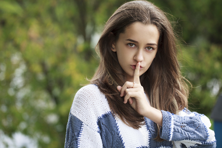 Young beautiful brunette woman has put forefinger to lips as sign of silence, against green summer gardenの写真素材
