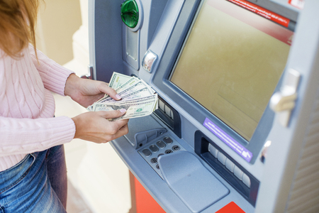 Young beautiful blonde woman is holding a cash dollars on the background of the ATM in a shopping centerの写真素材