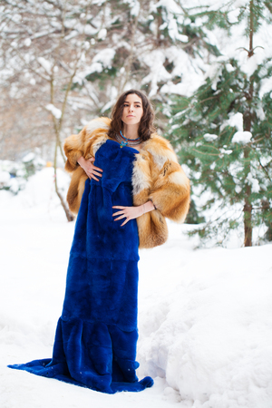 Young beautiful brunette girl in blue fur coat posing on winter park. Model wearing stylish warm clothes.の写真素材