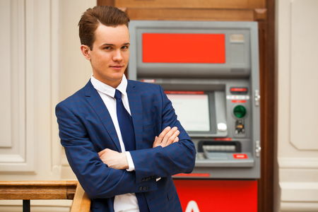 A young businessman in a blue suit stands against the background of an ATM in a shopping centerの写真素材