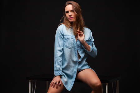 Portrait of a young sexy girl in a denim shirt and nude beautiful legs sitting on a table in a dark studioの写真素材