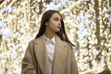 Portrait of a young girl in a beige coat against the backdrop of New Year's garlands. night view