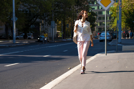 Portrait in full growth, young beautiful brunette woman in white blouse and pink pants walking on the street, summer outdoorsの写真素材