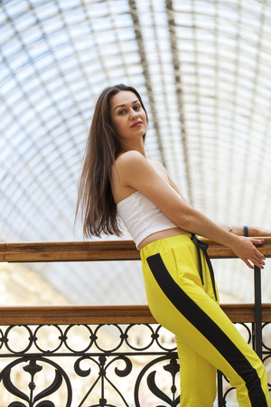 Portrait of a young beautiful girl in a white top and yellow sports pants posing in a mallの写真素材