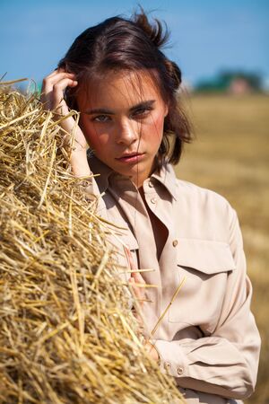 Young beautiful brunette woman in a beige dress posing on a background of haystacks in a cut fieldの写真素材