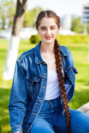 Young beautiful girl in a denim jacket sits on a bench in a summer park