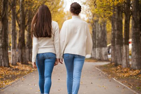 Back view, Two girlfriends in a white woolen sweater and blue jeans walk in the autumn parkの写真素材