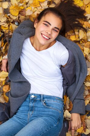 Top view portrait of a young beautiful girl in blue jeans and gray coat lies on autumn leaves in a parkの写真素材