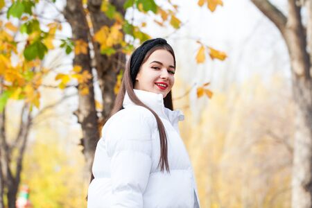 Close up portrait of a young beautiful woman in a white down jacket posing in autumn park