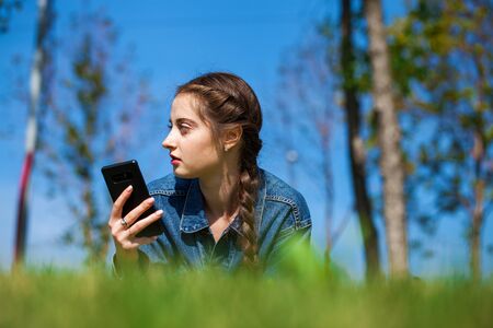 Close up portrait of beautiful young brunette woman in jeans jacket lying on the green grass, summer park outdoorsの写真素材