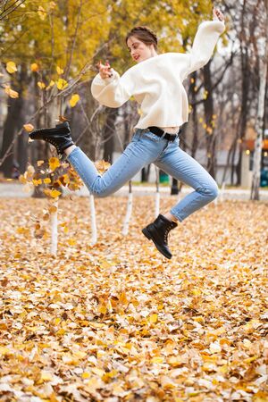 Portrait of a young beautiful girl in blue jeans and gwhite sweater sits on autumn leaves in a park