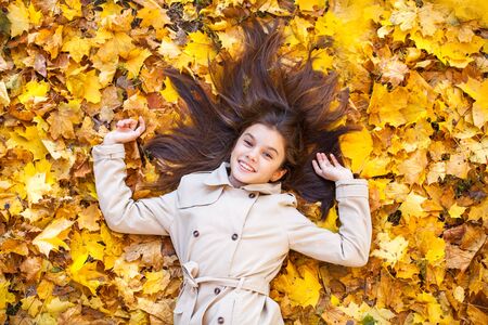 Young beautiful little girl in beige coat lying on yellow leaves, view from above, in the autumn...