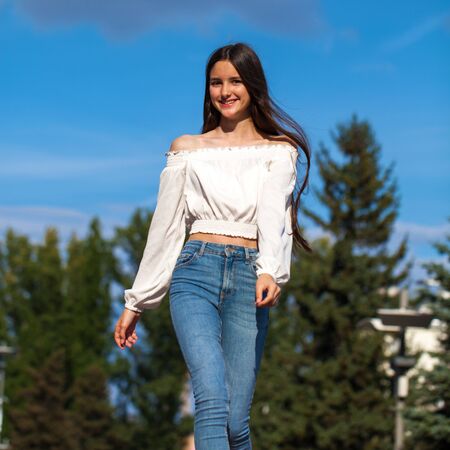 Portrait of young beautiful brunette woman in jeans and white blouse walking in summer streetの写真素材