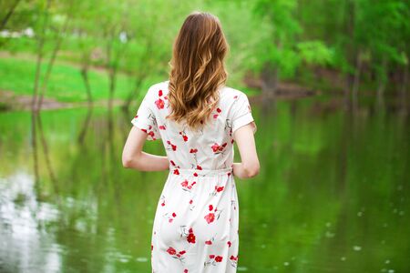 Hair back view. Close up young blonde woman in white dress posing by the lake