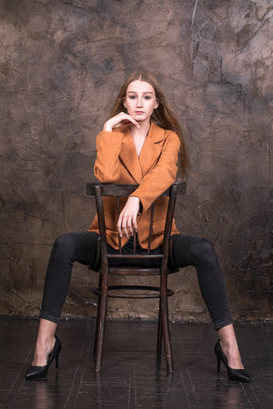 Young stylish girl in a suede orange jacket posing while sitting on a chair in the studioの写真素材