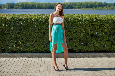 Full-length portrait of a young slender beautiful girl in a turquoise dress posing on the embankmentの写真素材