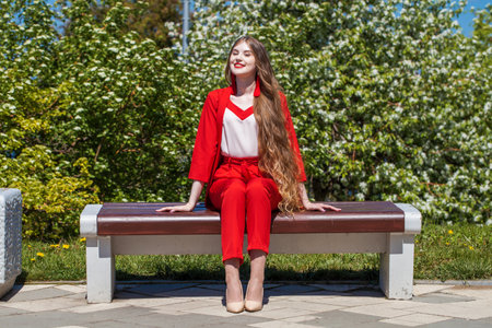 Full length portrait of a young blonde woman. A beautiful girl in a red bright suit sits on a bench in a spring parkの写真素材