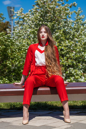Full length portrait of a young blonde woman. A beautiful girl in a red bright suit sits on a bench in a spring parkの写真素材