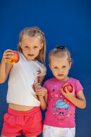 Two little girls posing with two ripe apples against blue wallの写真素材