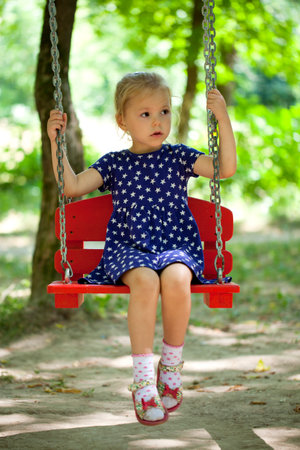 Adorable little girl having fun on a swing, against background of summer parkの写真素材