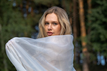 Young beautiful blonde girl posing with transparent fabric, summer park outdoorの写真素材