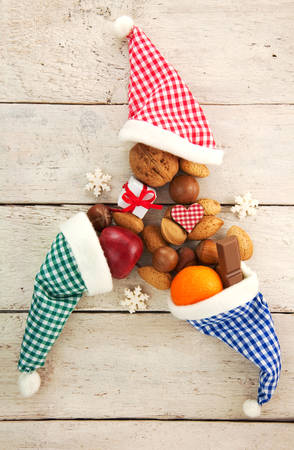 Three red, green, blue and white checkered Santa hats with candy, gift, nuts and Christmas decoration on a rustic wooden background for Christmas or St. Nicholasの写真素材