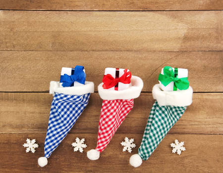 Three red, green, blue and white checkered Santa hats with gifts and Snowflakes on a rustic wooden background for Christmas or St. Nicholasの写真素材