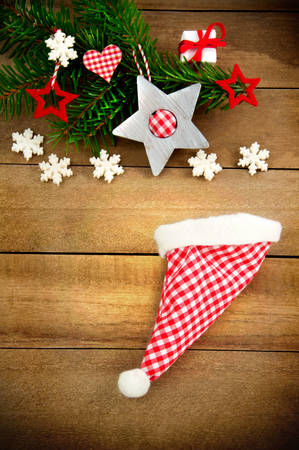 Rustic brown wooden background decorated with green fir branch, red and red and white checkered stars and santa hat, gift and snowflakes as christmas decoration in Country Styleの写真素材