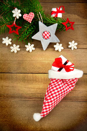 Rustic brown wooden background decorated with green fir branch, red and red and white checkered stars and santa hat, gift and snowflakes as christmas decoration in Country Styleの写真素材